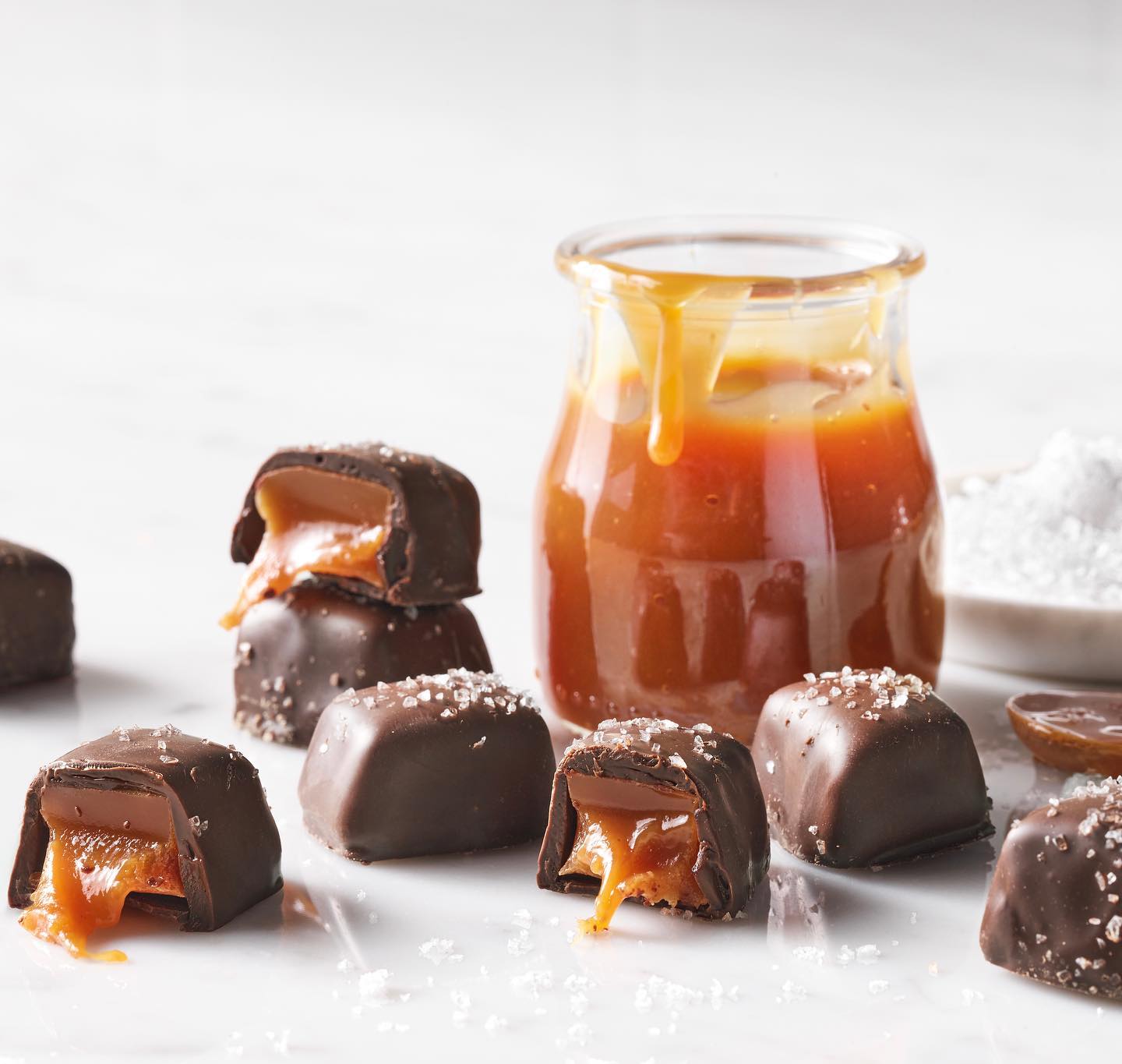 Chocolate-dipped salted caramel treats on a white surface with a glass container of caramel sauce.