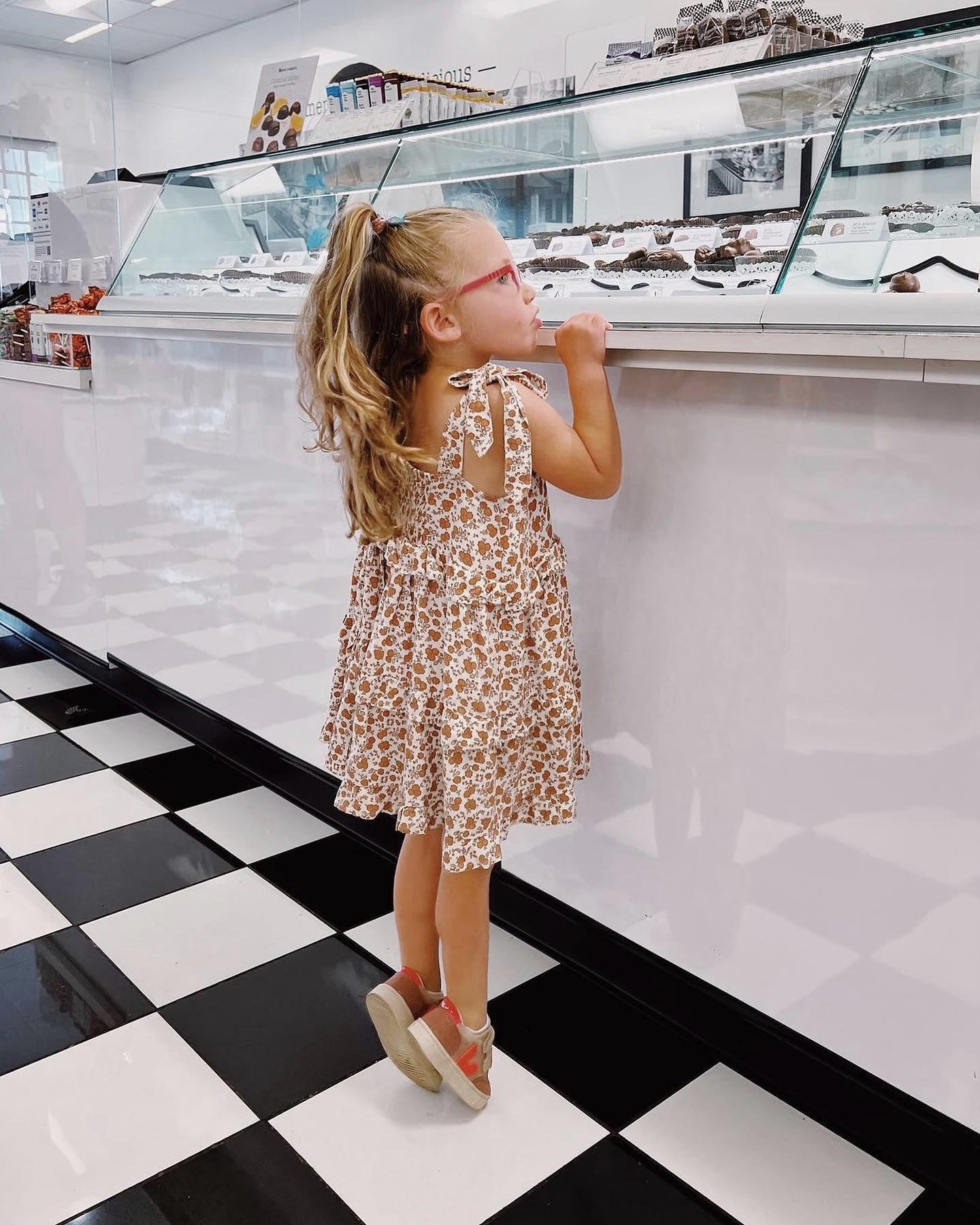 Young girl in a patterned dress standing on a checkered floor in a bakery.