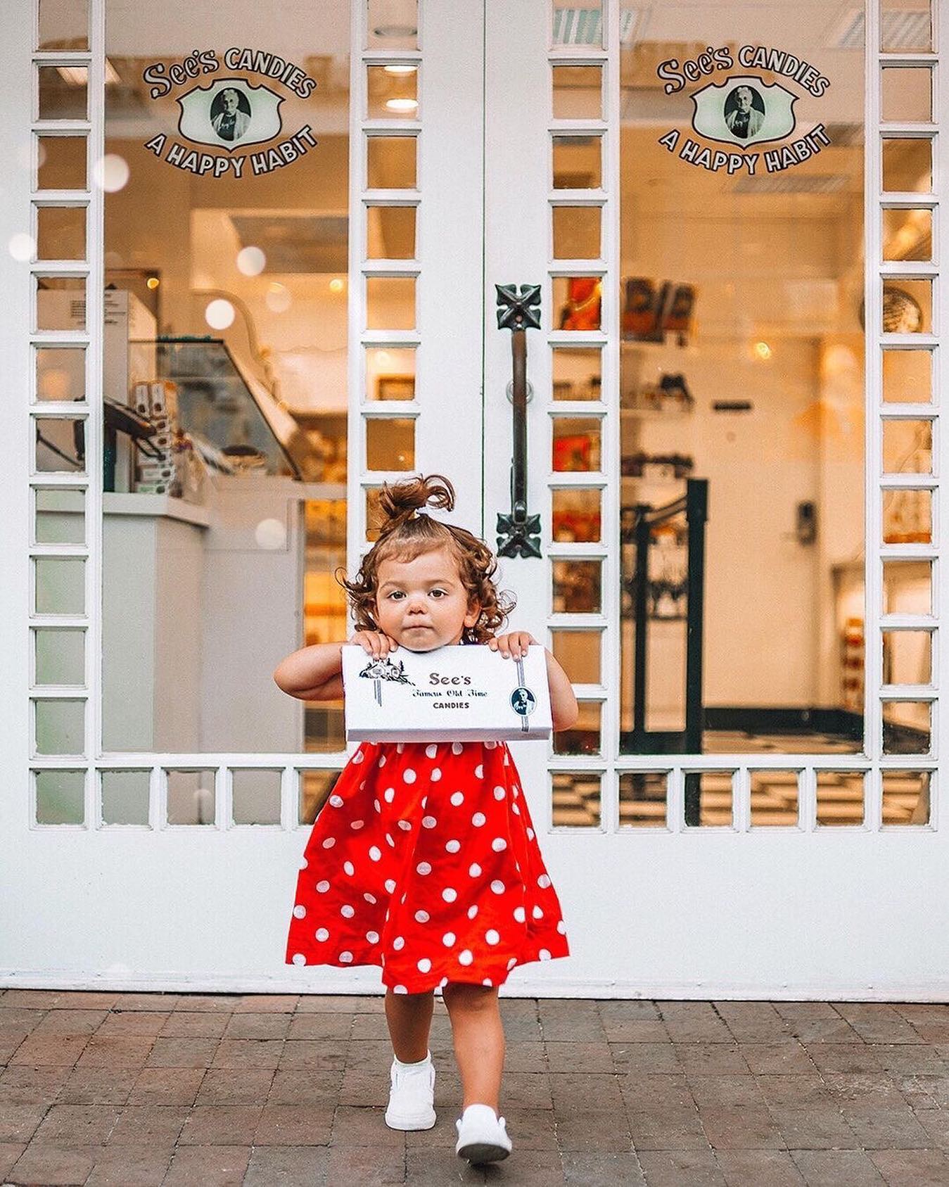 Child in a red polka dot dress holding a sign in front of a store entrance.