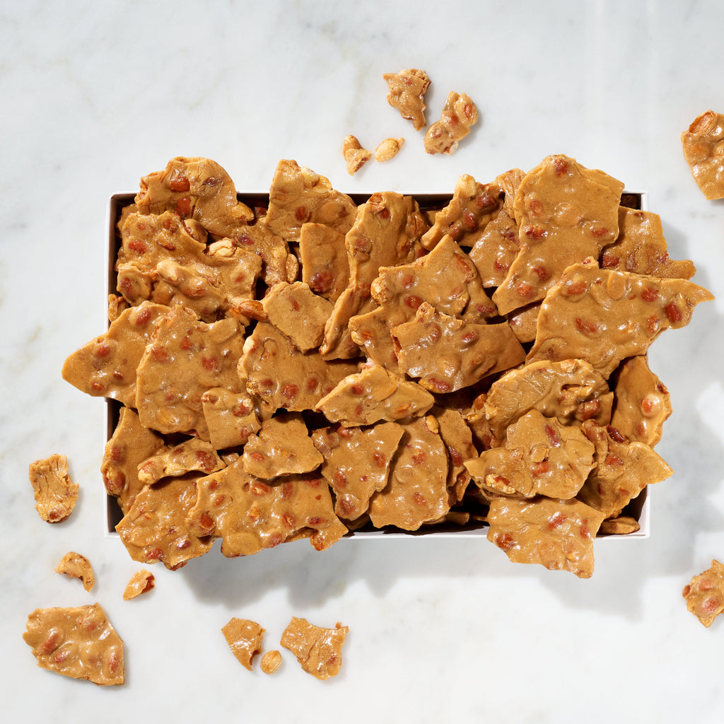 Peanut brittle pieces scattered on and around a dark rectangular box, photographed from above on a white marble surface.