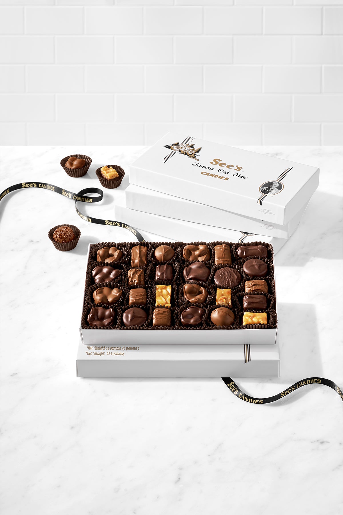 An open white gift box filled with nuts and chews chocolates sits on a white marble surface against a white subway-tile wall. A second closed white box is behind it, and a few chocolates are placed beside the open box.