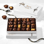 An open white gift box filled with assorted chocolates sits on a white marble surface against a white subway-tile wall. A second closed white box is behind it, and a few chocolates are placed beside the open box.