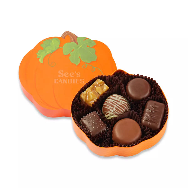 An orange pumpkin-shaped box of chocolates, shown open with the lid leaning behind it. The lid is decorated with a green pumpkin leaf design and the brand name 'See's CANDIES' is printed on it.