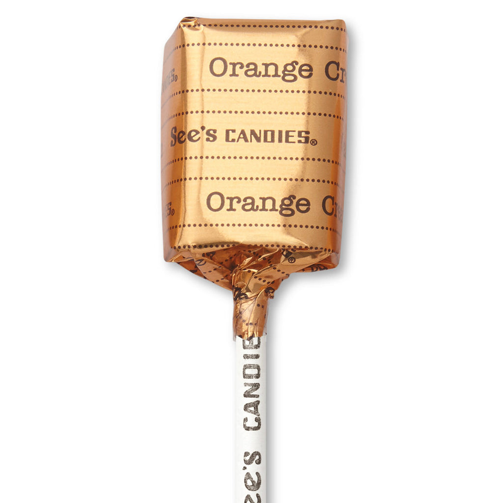 An individually wrapped lollipop from See's Candies in orange flavor, wrapped in metallic copper-orange foil with the brand name printed vertically on the stick and horizontally on the wrapper, set against a plain white background.