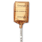 An individually wrapped lollipop from See's Candies in orange flavor, wrapped in metallic copper-orange foil with the brand name printed vertically on the stick and horizontally on the wrapper, set against a plain white background.